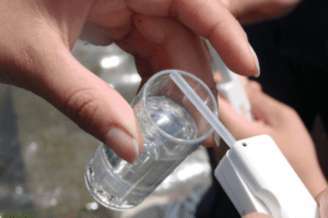 A close-up shows a sample of clear water being analyzed with a device.